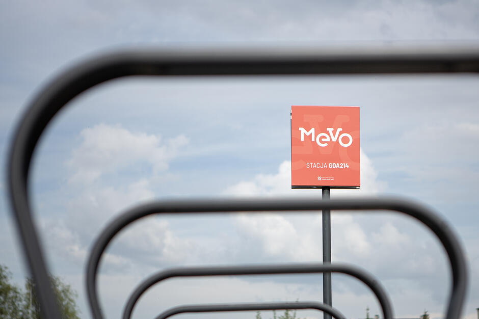 Zdjęcie przedstawia czerwoną tablicę informacyjną systemu roweru metropolitalnego Mevo z oznaczeniem stacji GDA214. Tablica umieszczona jest na słupku, a na pierwszym planie widać charakterystyczne, metalowe barierki służące do parkowania jednośladów.