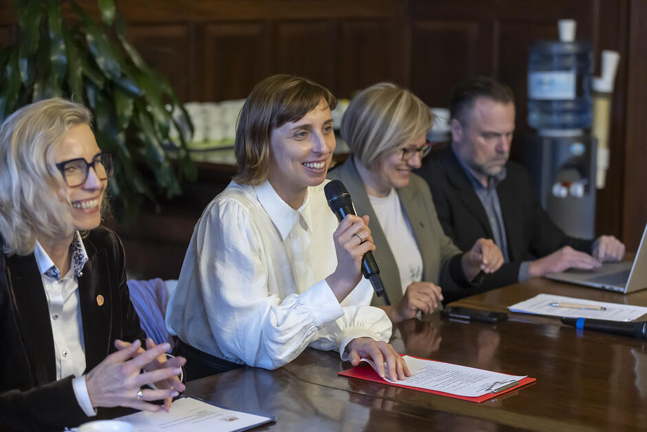 na zdjęciu trzy kobiety i jeden mężczyzna siedzą przy dużym stole, kobieta w środku mówi do mikrofonu