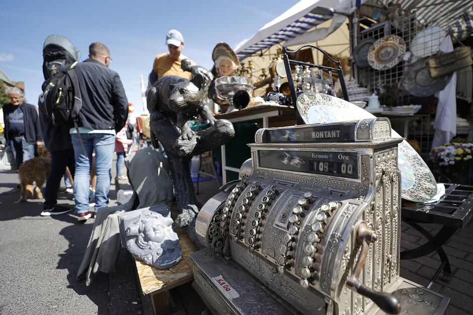 na zdjęciu po prawej wielka zabytkowa dawna kasa fiskalna w srebrnym kolorze, w tle stoiska jarmarkowe i przechodzący ludzie