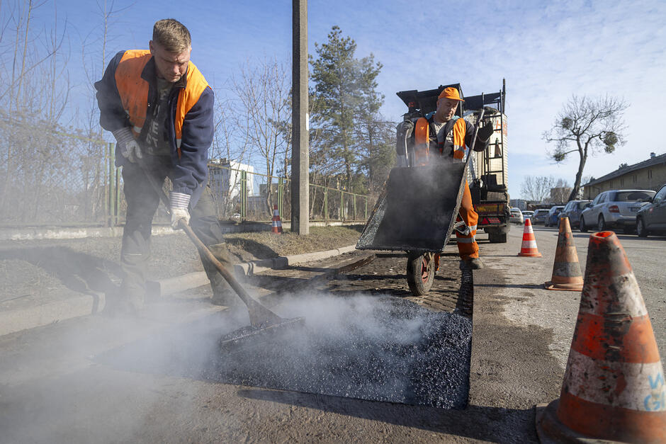 na zdjęciu dwóch mężczyzn w roboczych pomarańczowych kombinezonach łata dziurę w jezdni