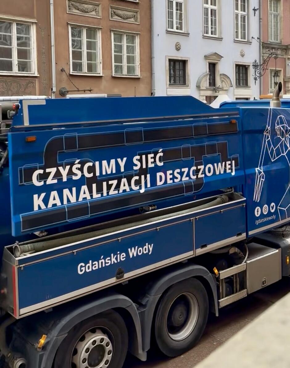 Zdjęcie przedstawia niebieski samochód ciężarowy należący do spółki Gdańskie Wody, który stoi na tle zabytkowych kamienic. Na boku pojazdu widnieje duży napis informujący, że służy on do czyszczenia sieci kanalizacji deszczowej.