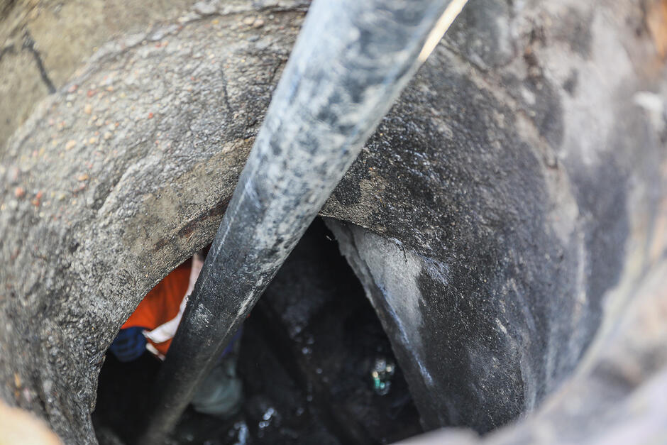 Widok z góry ukazuje wnętrze betonowej studzienki kanalizacyjnej, do której wprowadzona jest długa, ciemna rura. W głębi otworu dostrzec można fragmenty odzieży roboczej pracowników wykonujących czynności serwisowe.