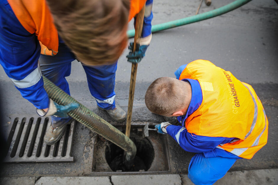 Dwóch pracowników spółki Gdańskie Wody w odblaskowych kamizelkach czyści studzienkę kanalizacyjną przy użyciu długiej rury ssącej i drążka. Jeden z mężczyzn klęczy przy otwartym włazie, podczas gdy drugi operuje sprzętem z góry, pilnując przebiegu prac serwisowych.
