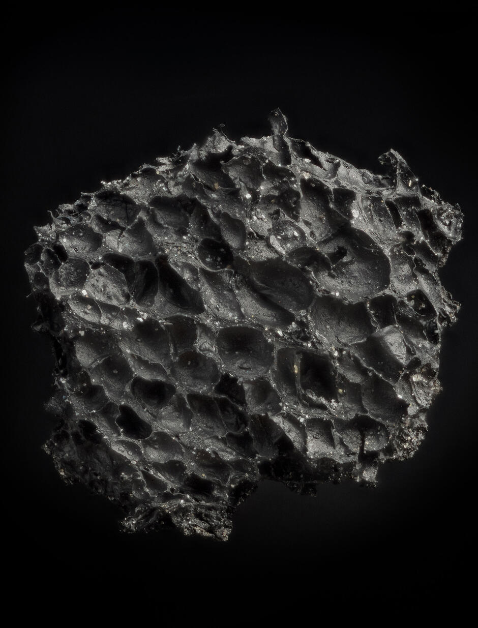 Zdjęcie przedstawia czarny, nieregularny obiekt o porowatej strukturze, który przypomina zastygłą lawę lub fragment meteorytu. Powierzchnia materiału jest pokryta licznymi zagłębieniami i pęcherzykami, nadającymi mu surowy, organiczny wygląd na ciemnym tle.
