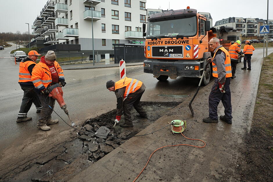 na zdjęciu drogowcy w pomarańczowych kamizelkach, dwóch rozkuwa asfalt młotami pneumatycznymi, w tle ciężarówka