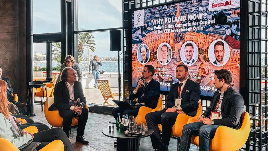 Czterech mężczyzn siedzi w fotelach na scenie - prowadzą dyskusję. Po lewej jest widownia przysłuchująca się tej rozmowie 