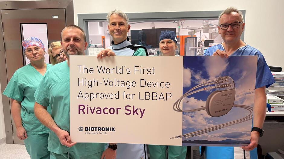 sześć osób stoi, patrzy się w obiektyw trzymając w ręce kartonową reklamę. Mają na sobie zielone i niebieskie ubrania, prawdopodobnie są lekarzami