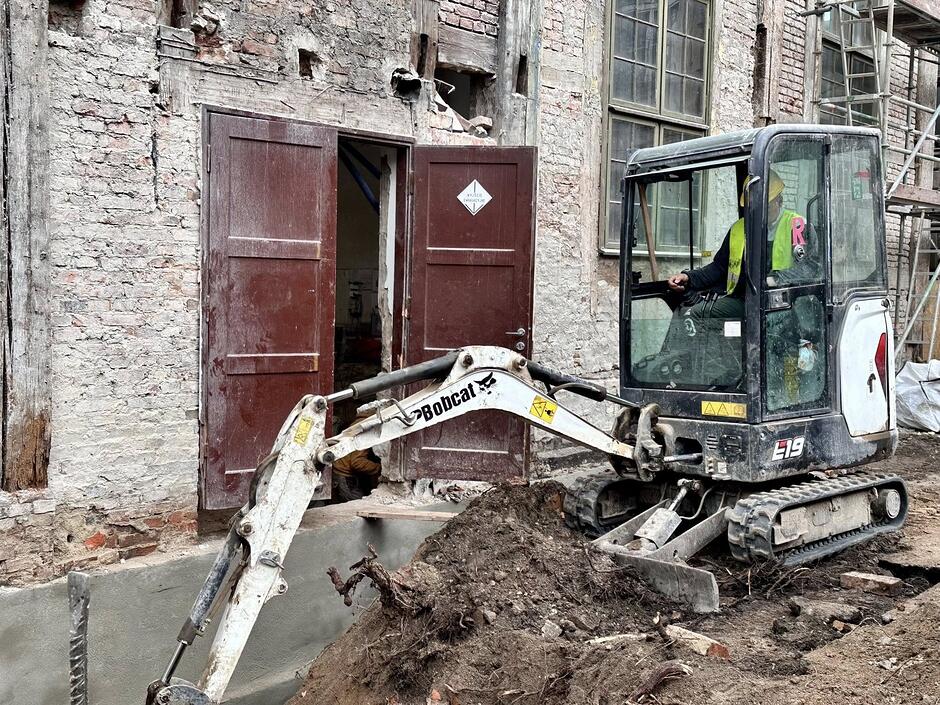 mała koparka z człowiekiem w środku, i rozkopany teren przed budynkiem