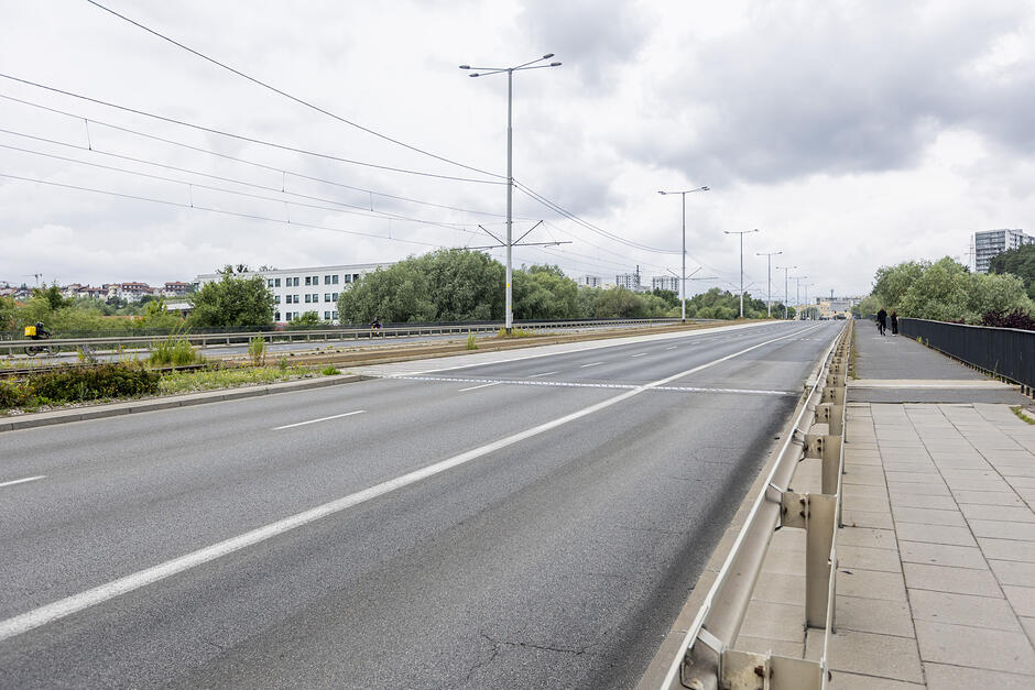 na pierwszym planie pusta jezdnia. Obok niej chodnik, a w tle budynki i zieleń