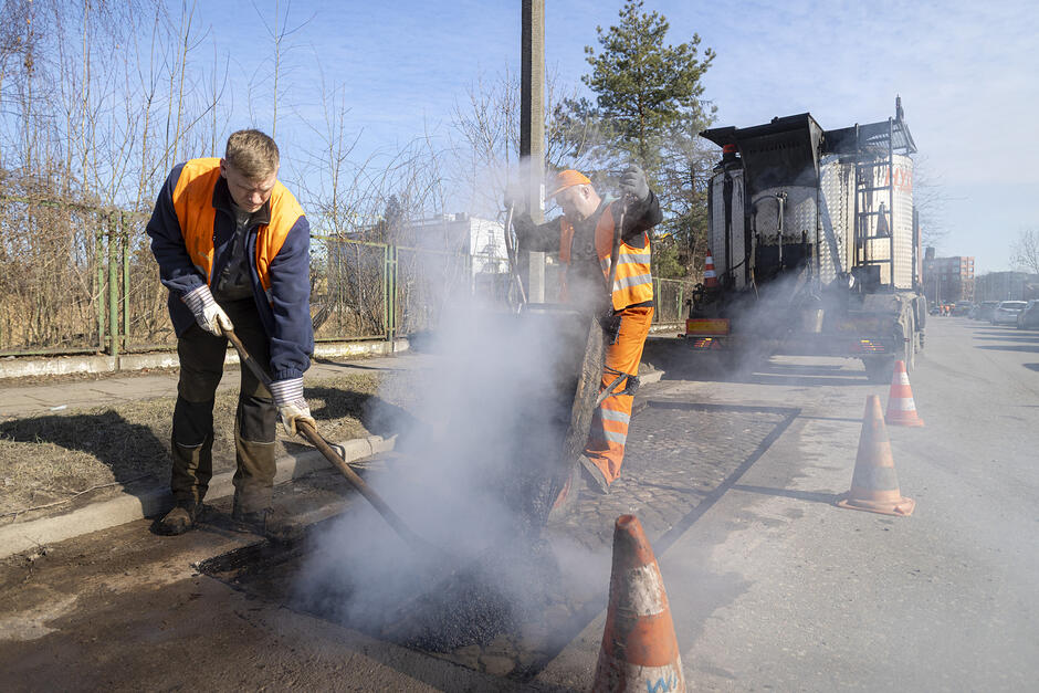 na zdjęciu drogowiec naprawiający nawierzchnię jezdni, widać zadymienie nad jezdnią a mężczyzna rozprowadza materiał łopatą