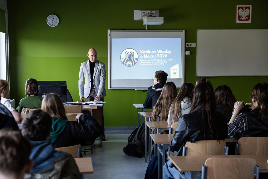 W sali w szkole siedzą uczniowie, widziani są od tyłu, z przodu stoi mężczyzna obok ekranu z napisem Konkurs Wiedzy o Morzu .