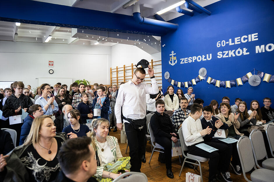 W sali gimnastycznej siedzą na krzesłach uczniowie i osoby dorosłe, środkiem idzie uczeń w białej koszuli, który ściąga z głowy czapkę, ludzie bija my brawo.