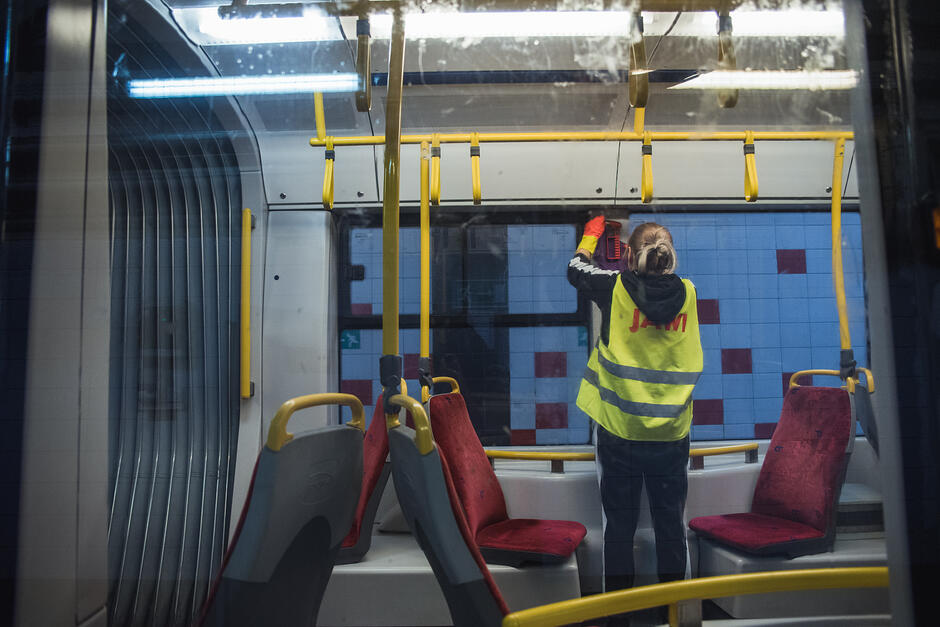 Zdjęcie przedstawia wnętrze nowoczesnego tramwaju lub autobusu, w którym osoba w żółtej kamizelce odblaskowej wykonuje prace porządkowe. W tle, przez szybę pojazdu, widoczna jest ściana stacji metra wyłożona charakterystycznymi niebieskimi i czerwonymi kafelkami.