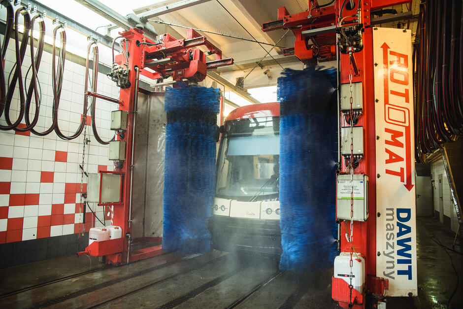 Zdjęcie przedstawia nowoczesny tramwaj znajdujący się wewnątrz automatycznej myjni wielkogabarytowej. Pojazd jest czyszczony przez pionowe, niebieskie szczotki obrotowe zamontowane na czerwonej, przemysłowej konstrukcji marki Rotomat. 