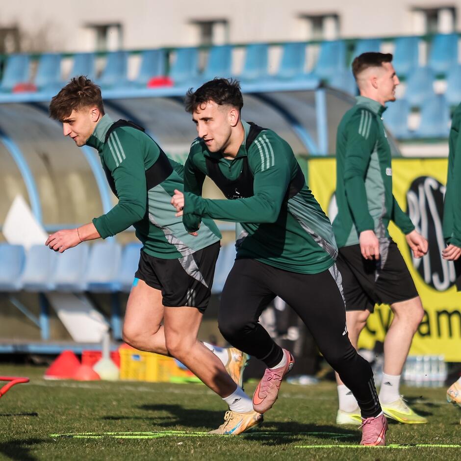 Dwaj po lewej rozpoczynają sprint na murawie. Trzeci - po prawej - stoi odwrócony do nich bokiem