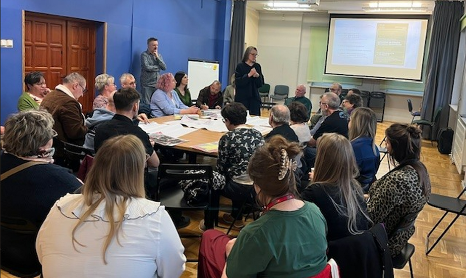 niewielka sala z niebieskimi ścianami, po środku stół wokół którego siedzi około dwudziestu osób