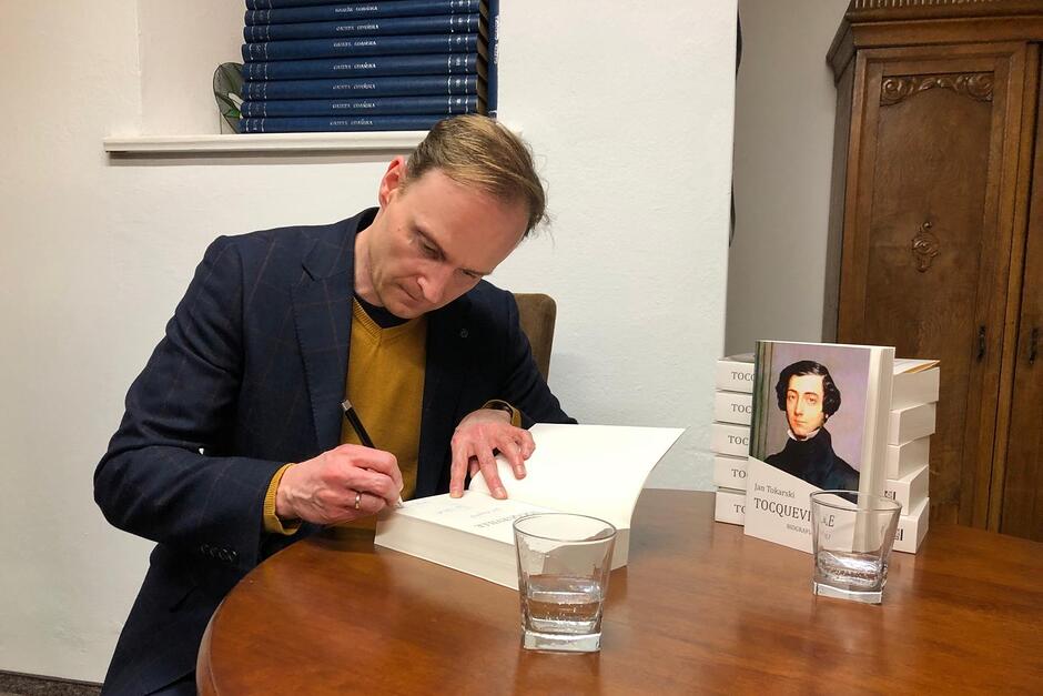 Jan Tokarski siedzi przy stole po lewej, do książki wpisuje dedykację dla uczestnika spotkania autorskiego. Po prawej na stole leżą i stoją egzemplarze książki o Toquevillu  