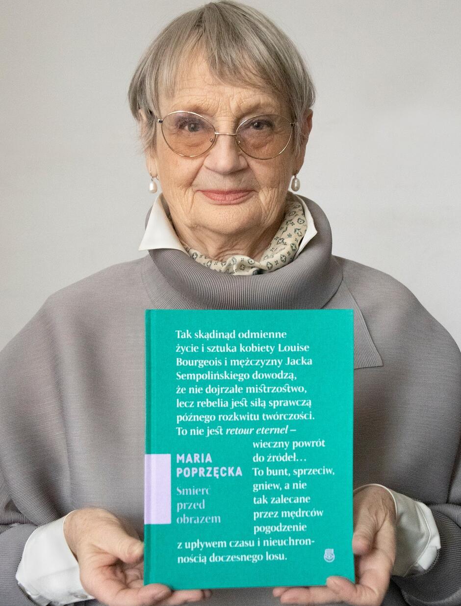 Starsza kobieta w okularach, w rękach na wysokości trzyma książkę, która jest skierowana okładką do widza