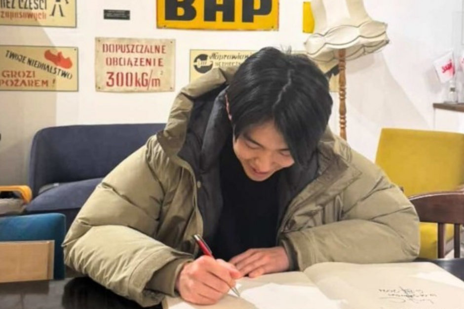 mężczyzna z długimi włosami siedzi przy stole i z uśmiechem na twarzy pisze coś w księdze. Ma na sobie kurtkę, z tyłu jest szara sofa. Wygląda jakby siedział w ciasnym pomieszczeniu