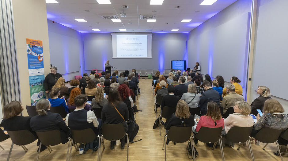 Na sali konferencyjnej siedzą ludzie na krzesłach, zwróceni w stronę mównicy, na której przemawia kobieta.