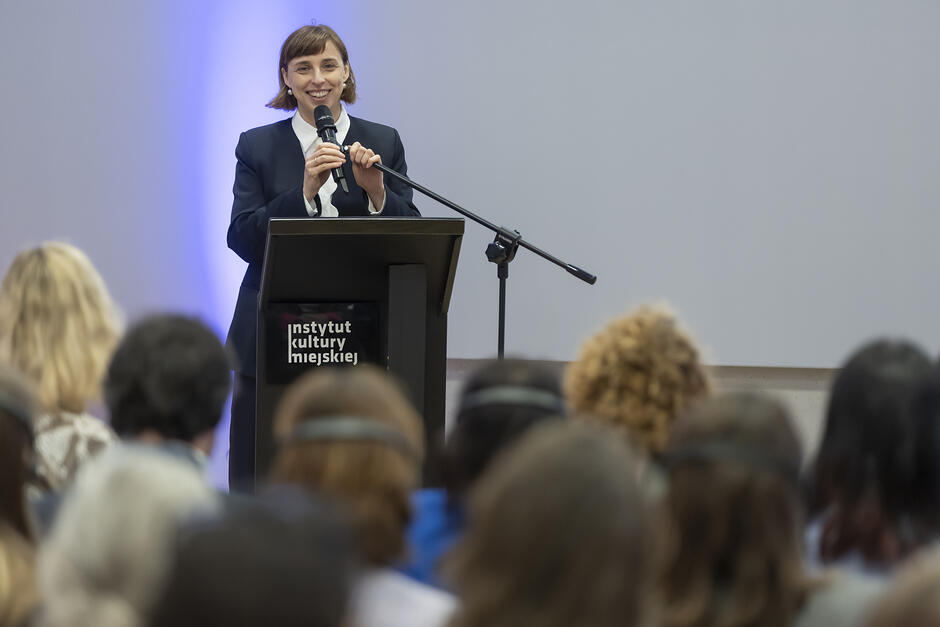 Kobieta przemawia na mównicy w sali konferencyjnej, przed nią siedzą na krzesłach ludzie.