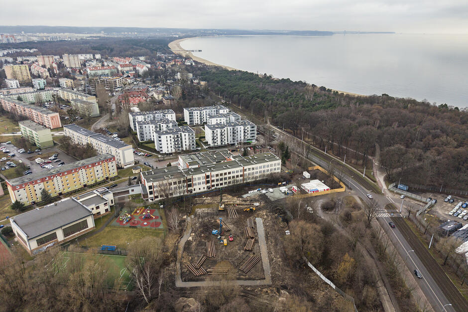 zdjęcie z drona, widać plac budowy, w tle zabudowę mieszkaniową i duży budynek szkoły, w tle wody zatoki gdańskiej