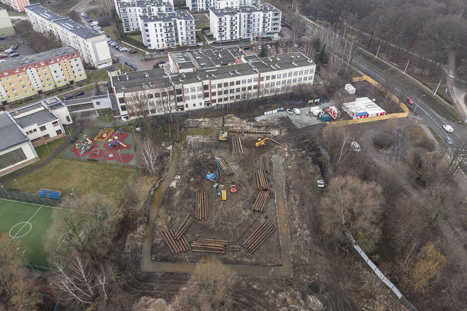 zdjęcie z drona, widać plac budowy i żółte koparki, obok budynek szkolny