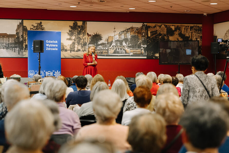 Spotkanie w Bibliotece Manhattan. Na scenie Laura Łącz opowiada swoją historię opieki nad schorowaną mamą. Tłum uczestników wsłuchuje się w wystąpienie artystyki.