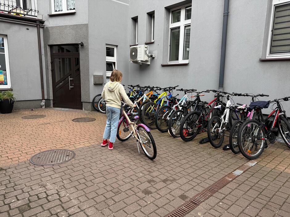 Zdjęcie przedstawia dziecko na dziedzińcu szkoły lub budynku mieszkalnego, które stawia swój rower obok długiego rzędu jednośladów przypiętych do stojaka. W tle widoczna jest szara elewacja budynku z oknami, drzwiami wejściowymi oraz zamontowaną jednostką klimatyzacji. 