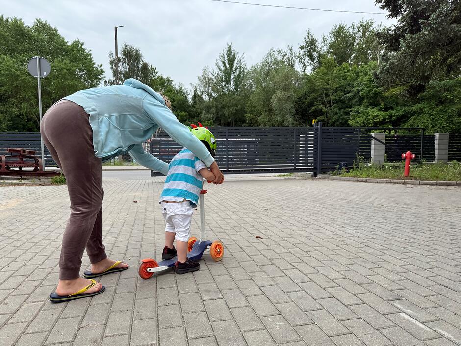 Na zdjęciu widać kobietę pomagającą małemu dziecku w kasku utrzymać równowagę na kolorowej hulajnodze. Scena rozgrywa się na wyłożonym kostką podjeździe, w tle którego znajduje się ciemne ogrodzenie oraz gęsta zieleń drzew.