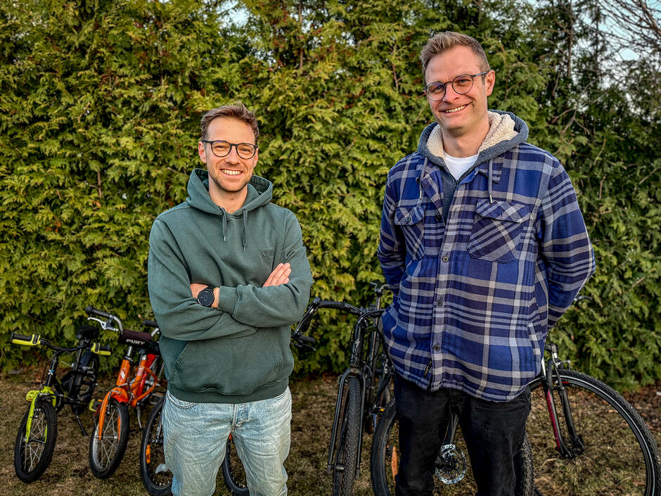 Dwaj uśmiechnięci mężczyźni w okularach pozują na tle gęstego żywopłotu z tui, otoczeni kilkoma rowerami o różnych rozmiarach. Jeden z nich ma na sobie zieloną bluzę z kapturem i skrzyżowane ramiona, natomiast drugi ubrany jest w grubą, niebieską koszulę w kratę.