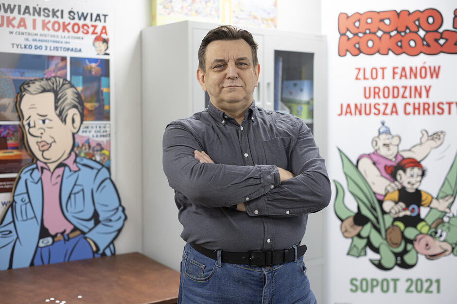 Mężczyzna w szarej koszuli stoi z założonymi rękami na tle plakatów i dekoracji związanych z komiksem „Kajko i Kokosz”. Obok niego widać dużą makietę przedstawiającą rysunkową postać oraz baner informujący o zlocie fanów w Sopocie. 