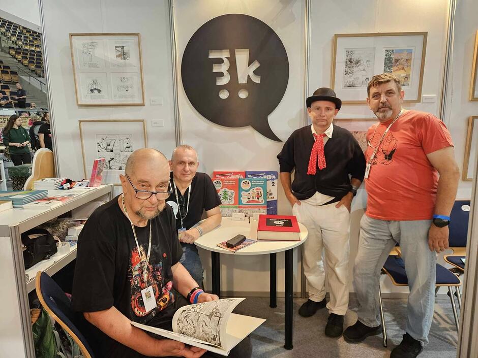 Na zdjęciu widać czterech mężczyzn pozujących przy stoisku na festiwalu komiksu, otoczonych planszami z grafikami i albumami. Jeden z nich siedzi na pierwszym planie, przeglądając otwartą książkę, podczas gdy pozostali patrzą w stronę obiektywu.