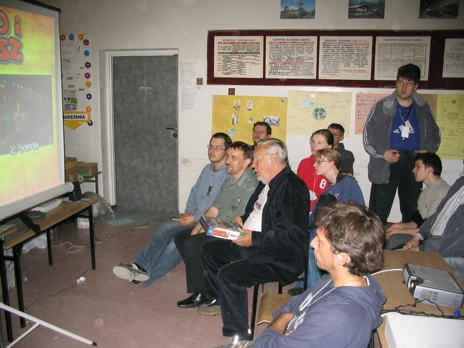 Grupa osób w różnym wieku siedzi w sali konferencyjnej lub klasie, uważnie oglądając prezentację wyświetlaną na dużym ekranie. W centralnym punkcie siedzi starszy mężczyzna trzymający pudełko, prawdopodobnie grę planszową, podczas gdy w tle widać tablice z informacjami i plakatami.