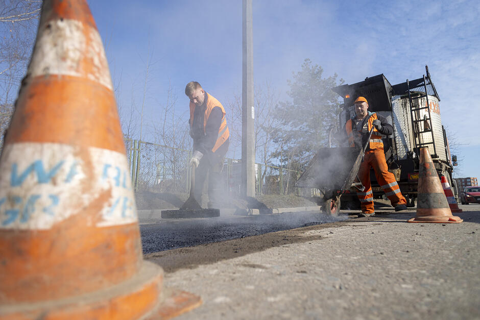 na pierwszym planie po lewej pomarańczowy pachołek drogowy ustawiony na jezdni, w tle dwóch robotników naprawia nawierzchnię jezdni, widać też duży jasny samochód