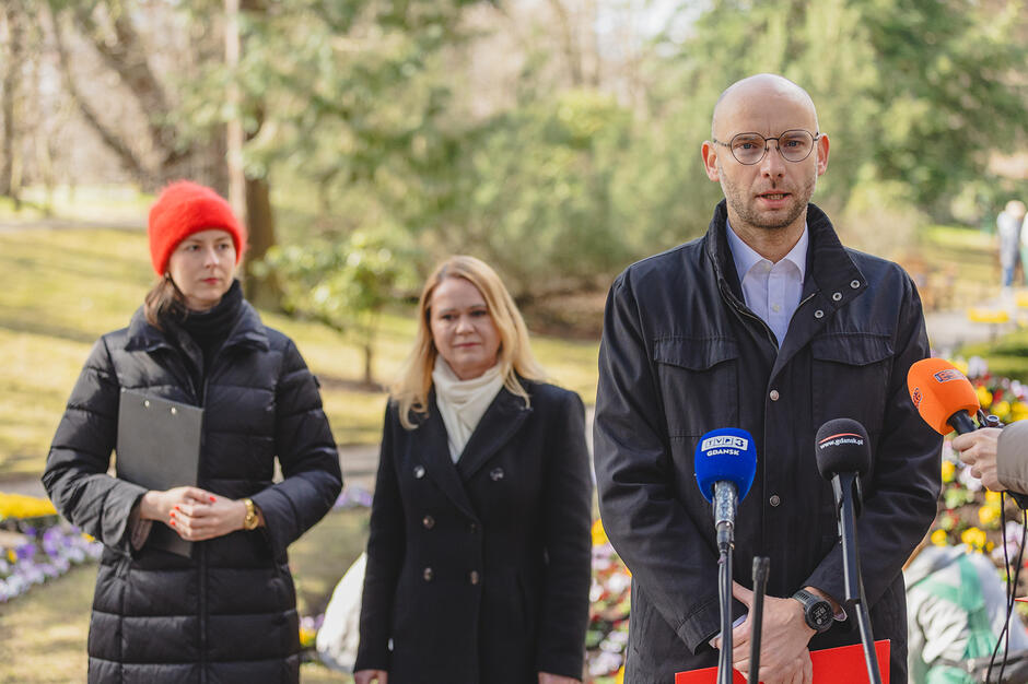 Mężczyzna mówi do mikrofonów w parku, za nim stoją dwie kobiety, w tle drzewa.