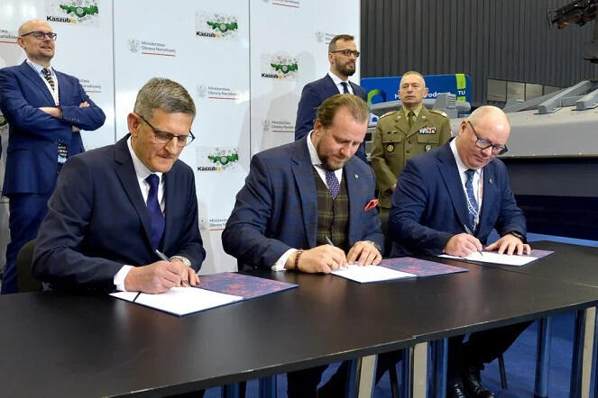 grupa trzech mężczyzn ubranych w garnitury siedzi przy stole i podpisuje dokumenty. W tle stoją trzy kolejne osoby