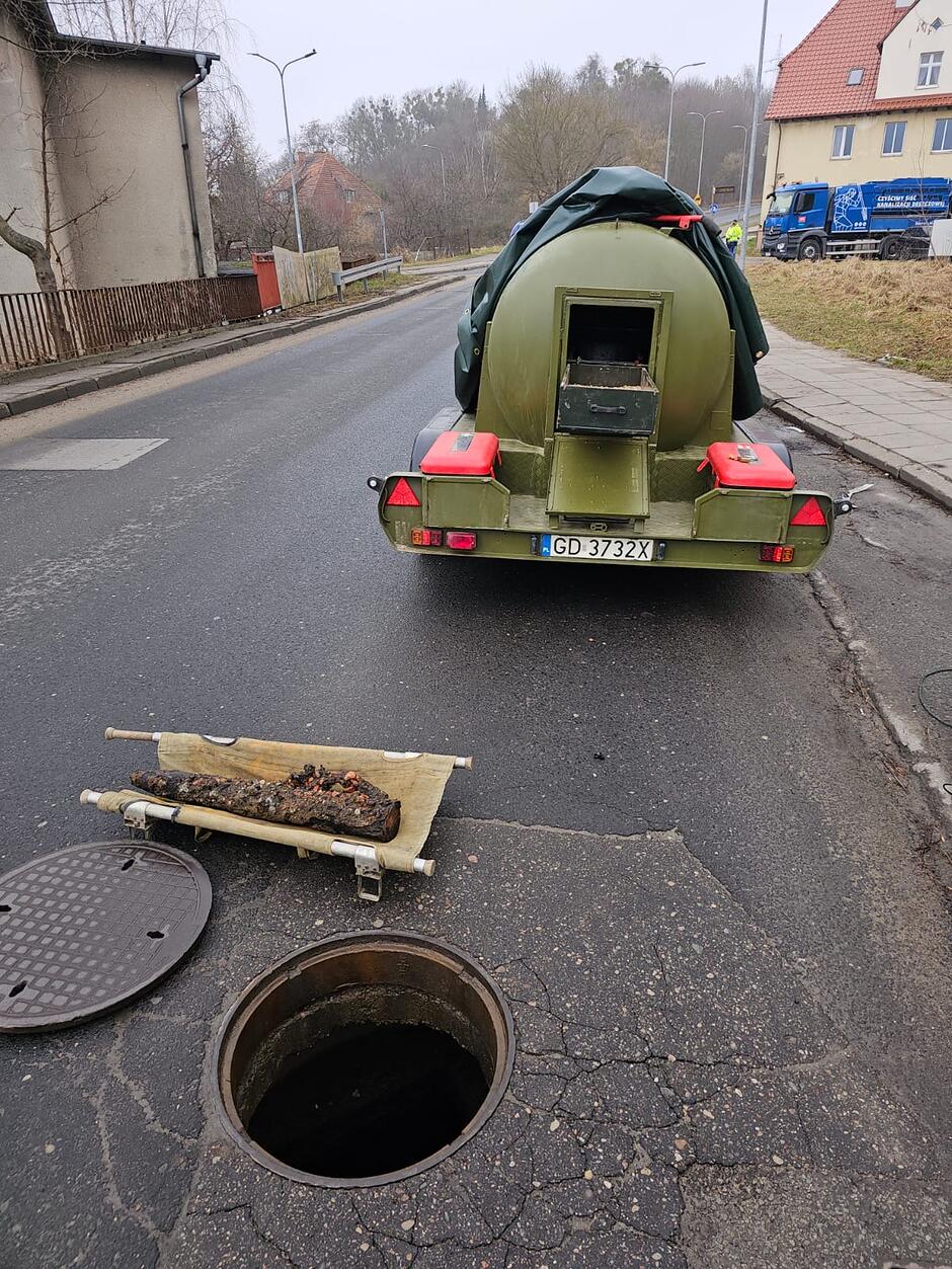Na zdjęciu widać interwencję patrolu saperskiego, który przy otwartym włazie kanalizacyjnym ustawił specjalistyczną przyczepę do bezpiecznego transportu niewybuchów. Na noszach leży wydobyty z kanału, silnie skorodowany przedmiot, który kształtem przypomina pocisk artyleryjski lub inną niebezpieczną pozostałość z czasów wojny.