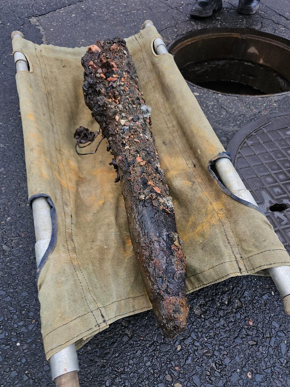 Na wojskowych noszach leży wydobyty z kanału, podłużny i silnie skorodowany przedmiot, który jest oblepiony warstwą ziemi i gruzu. Znalezisko przypomina pocisk artyleryjski lub inny niewybuch, a obok niego widoczny jest otwarty właz kanalizacyjny na asfaltowej drodze.
