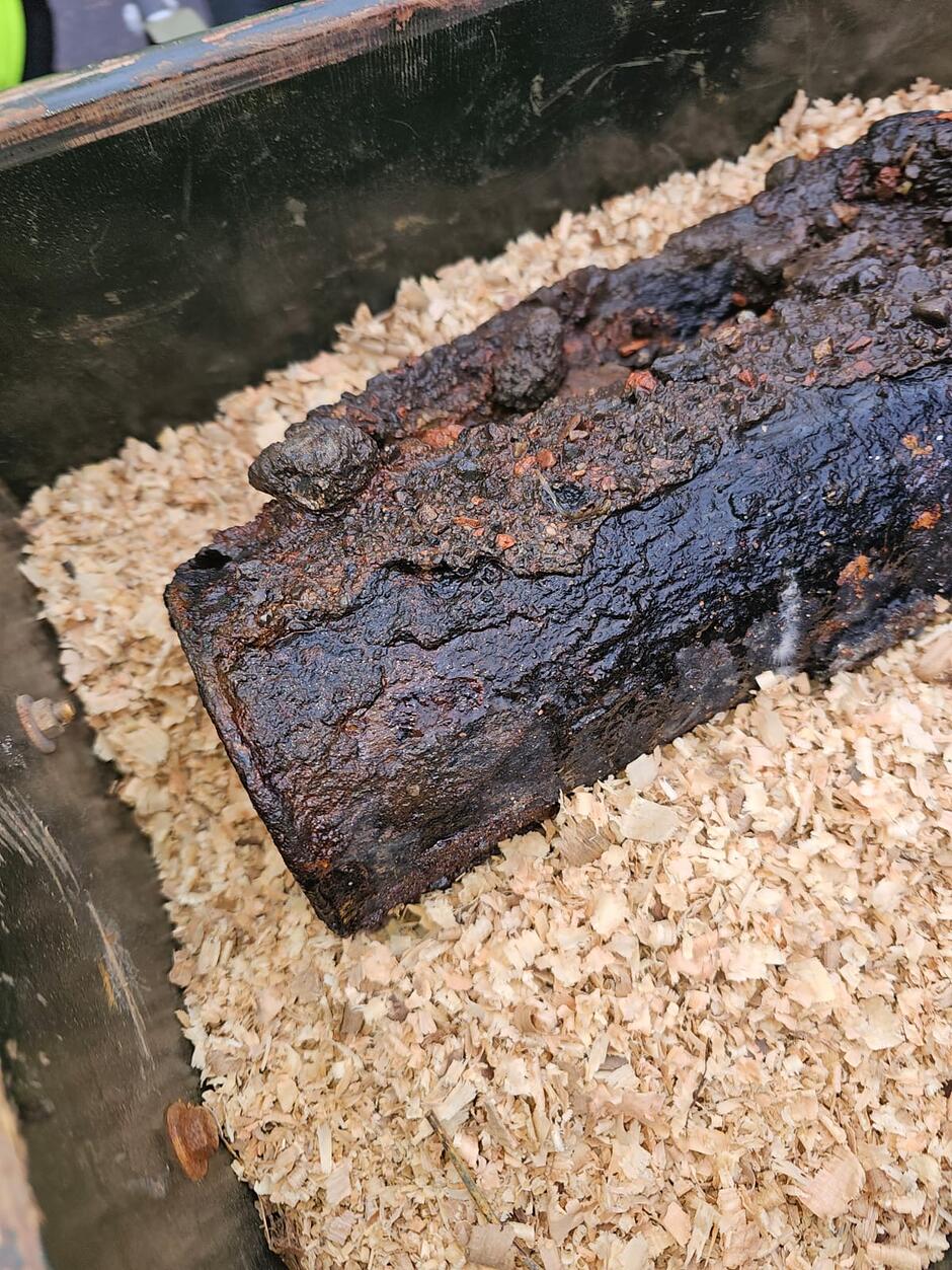 Zdjęcie przedstawia podłużny, silnie skorodowany i zwęglony przedmiot o nieregularnej fakturze, przypominający stary niewybuch lub fragment metalowej konstrukcji. Znalezisko spoczywa w drewnianej skrzyni wypełnionej jasnymi trocinami, które mają zapewnić mu stabilizację podczas transportu.