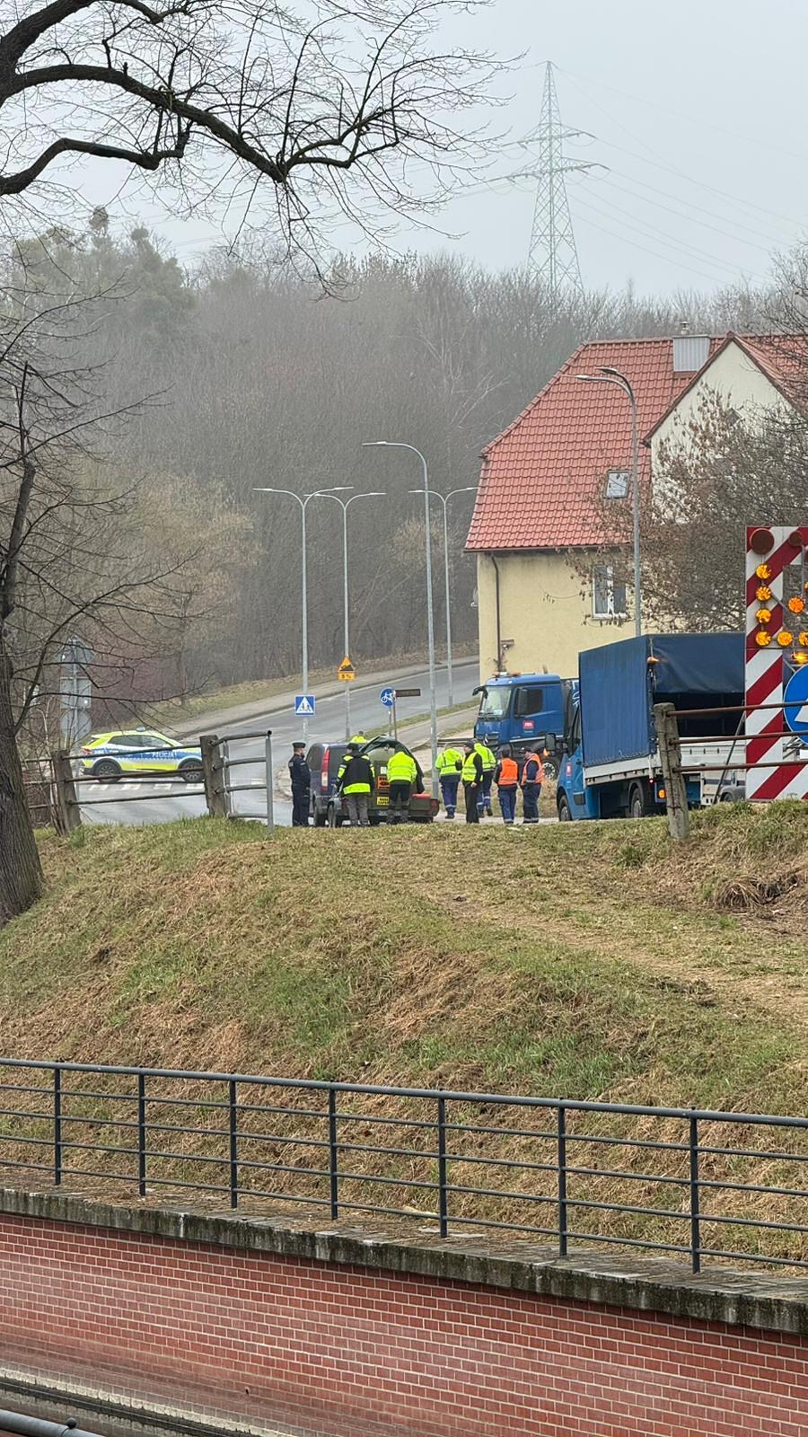 Na zdjęciu widać grupę służb mundurowych i technicznych pracujących przy drodze obok zaparkowanych samochodów ciężarowych oraz radiowozu. Całe zdarzenie ma miejsce na wzniesieniu w pobliżu budynków mieszkalnych i linii energetycznej, w pochmurny, bezlistny dzień.