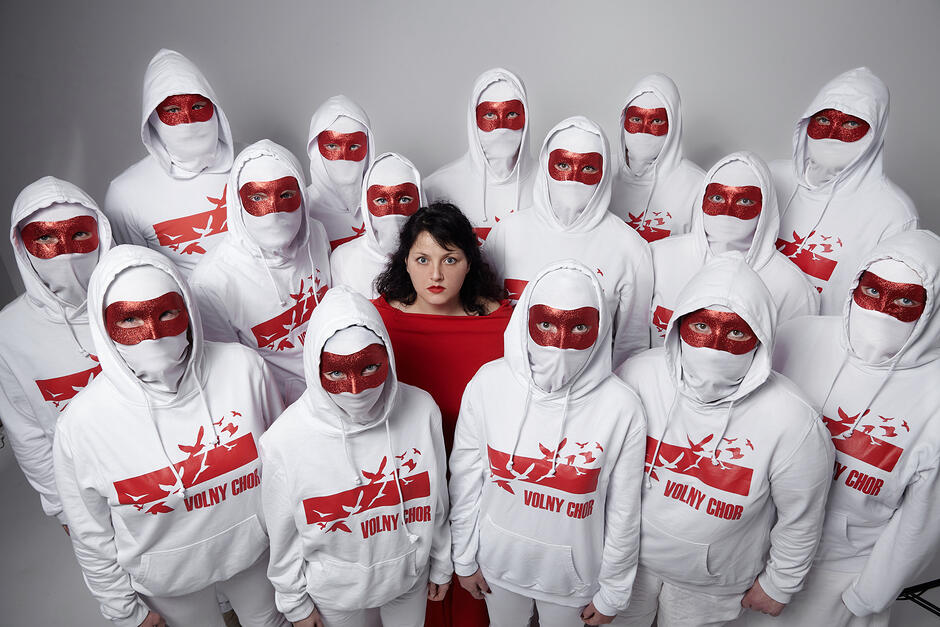 Zdjęcie przedstawia grupę osób w białych bluzach z kapturem i białych maskach z czerwonymi, błyszczącymi elementami wokół oczu. W centrum stoi kobieta w kontrastującej, czerwonej sukni, a na bluzach pozostałych osób widnieje czerwony pas z ptakami oraz napis „VOLNY CHOR”. 