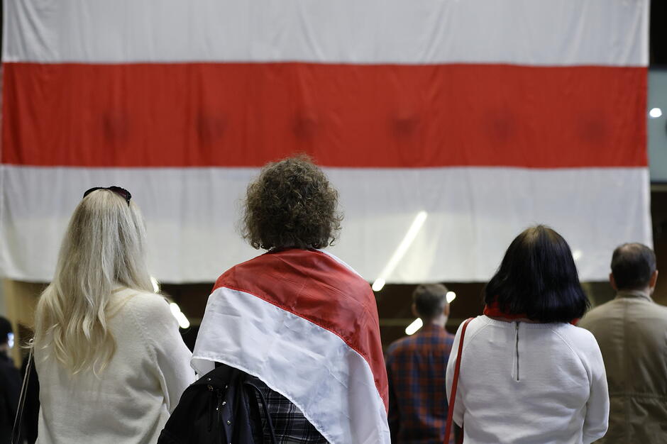 Zdjęcie przedstawia grupę ludzi stojących tyłem, którzy patrzą na dużą, biało-czerwono-białą flagę narodową Białorusi. Jedna z osób ma mniejszą flagę w tych samych barwach zarzuconą na ramiona, co sugeruje udział w wydarzeniu o charakterze patriotycznym lub solidarnościowym.