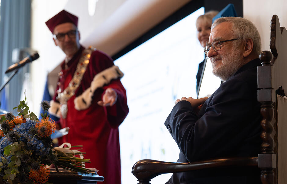 Na zdjęciu widać uroczystość akademicką, podczas której mężczyzna w ciemnym garniturze siedzi na ozdobnym krześle i uśmiecha się w stronę kamery. W tle stoi inna osoba ubrana w tradycyjną, czerwoną togę rektorską z ozdobnym łańcuchem, a na pierwszym planie znajduje się kolorowy bukiet kwiatów.