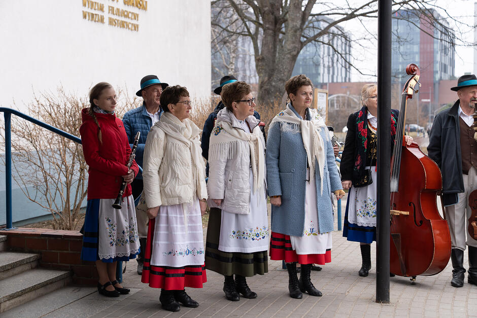 Na zdjęciu widać grupę osób w strojach ludowych, które stoją przed budynkiem Uniwersytetu Gdańskiego, trzymając instrumenty muzyczne, takie jak klarnet i kontrabas. Kobiety mają na sobie haftowane spódnice, białe chusty i kolorowe okrycia wierzchnie, co sugeruje oprawę artystyczną lokalnego wydarzenia.