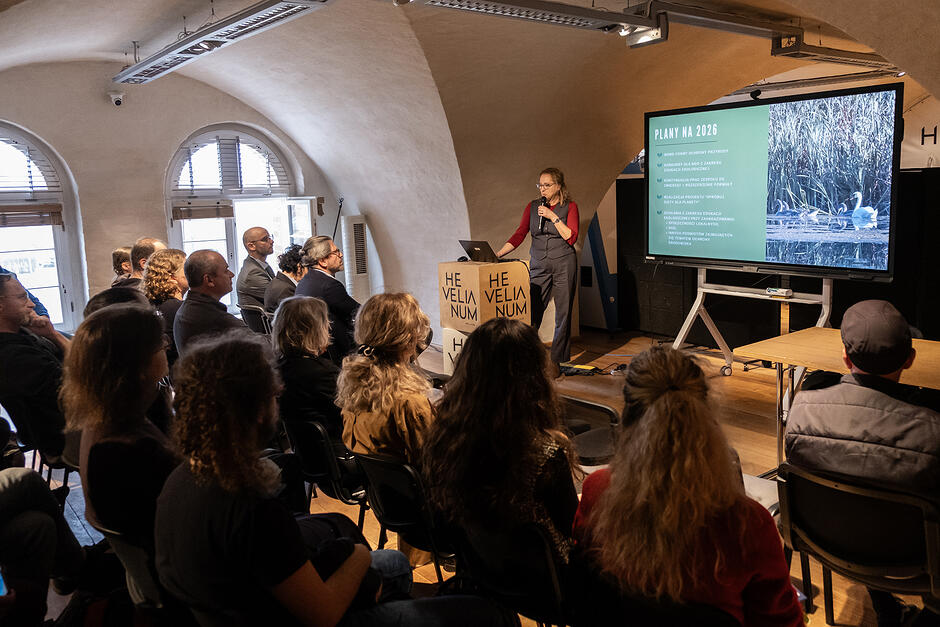 W sali konferencyjnej w starym budynku z łukowymi sklepieniami siedzą ludzie na krzesłach. Przed nimi stoi kobieta na mównicy i przemawia. Obok niej ekran z prezentacją.