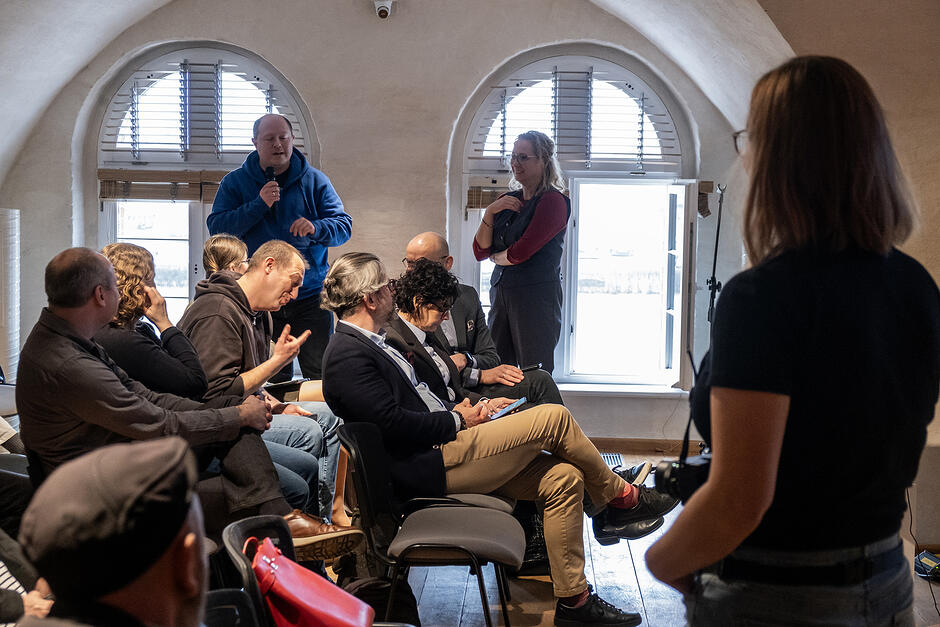 Kobieta i mężczyzna stoją w sali konferencyjnej w starym budynku z łukowymi oknami i sklepieniami. Mężczyzna mówi do mikrofonu. Wokół siedzą słuchacze na krzesłach.