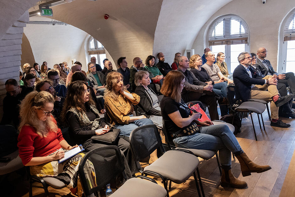 W sali konferencyjnej w starym budynku z łukowymi sklepieniami siedzą na krzesłach słuchacze konferencji.