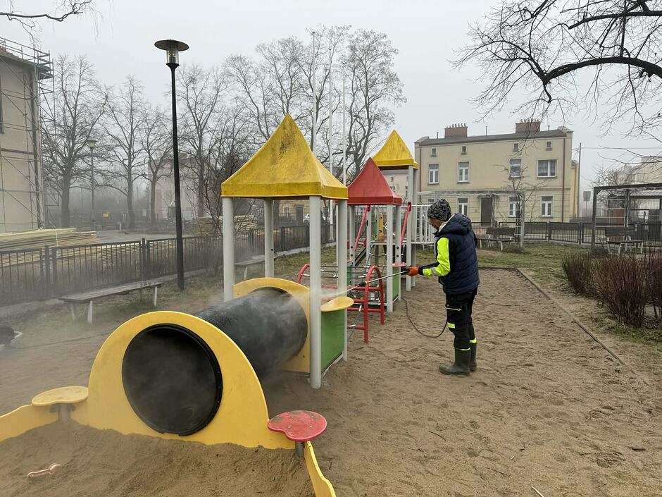 11na zdjęciu pracownik w czarnym stroju, myje za pomocą wody z węża urządzenie na palcu zabaw przypominające lokomotywę