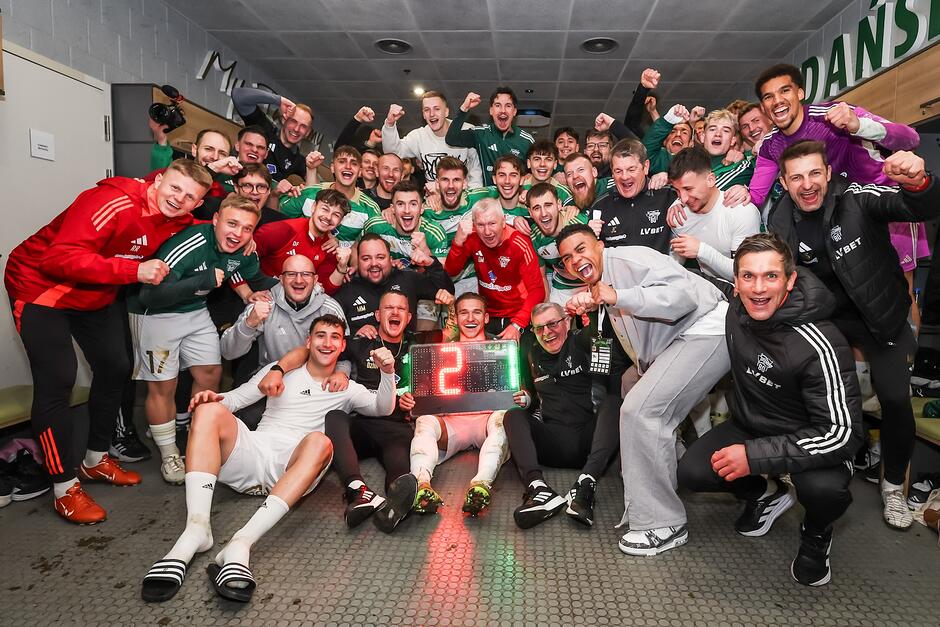 Zbiorowa radość około 30 mężczyzn w dresach lub strojach piłkarskich. Pozują do zdjęcia w szatni, z tablicą świetlną 